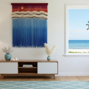 May include: A woven wall hanging with a gradient of colors from red to blue, depicting a sun and waves. Below, a wooden cabinet with white doors and a blue rug. A floor lamp and a window with a view of the ocean complete the scene.