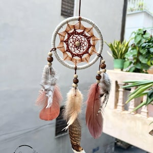 Op de afbeelding: Een witte droomvanger met bruine en beige geweven details, met drie veren in tinten roze, beige en bruin. De droomvanger hangt aan een bruin koord.