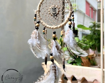 May include: A beige dreamcatcher with a woven web design, adorned with white and brown feathers, wooden beads, and a small owl charm. The dreamcatcher is suspended by a natural fiber cord, creating a bohemian aesthetic.