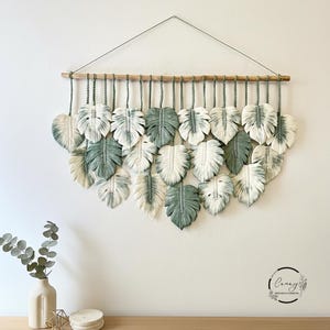Pode incluir: Uma tapeçaria de parede em macramé com desenhos em forma de folha em tons de branco e verde. As folhas são suspensas de um pino de madeira natural com cordas verdes. Decoração de parede.