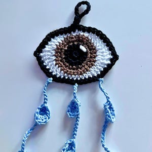 Op de afbeelding: Een gehaakt oog met een bruine iris en een zwarte pupil, met blauwe gehaakte traantjes die eronder hangen.