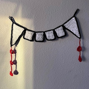 May include: A white and black crocheted banner with a jagged tooth design. Red crocheted strands hang from the ends of the banner.