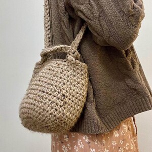 May include: A beige crocheted tote bag with a brown sweater and a floral skirt in the background.