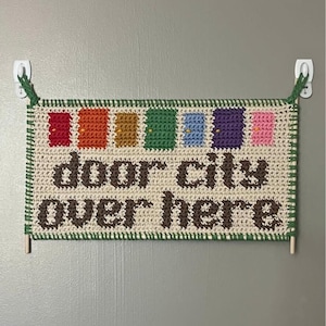 Pode incluir: Uma tapeçaria de parede de crochê com o texto "door city over here". A faixa apresenta uma série de desenhos de portas coloridas em vermelho, laranja, amarelo, verde, azul, roxo e rosa. A borda é verde e o texto é marrom.