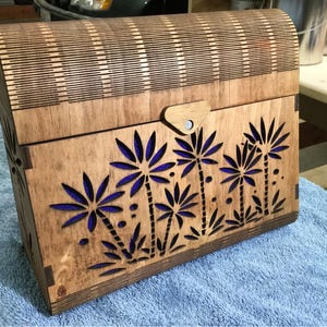Peut inclure: Une boîte en bois avec un couvercle à charnières et un loquet. La boîte est décorée d'un motif de palmiers et de feuilles découpés au laser dans des tons de bleu et de brun.
