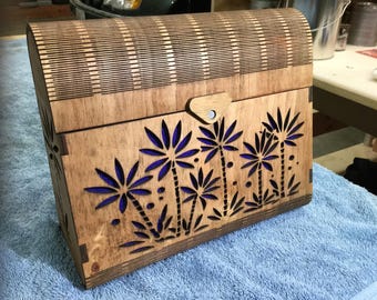 Puede incluir: Una caja de madera con tapa abatible y cierre. La caja está decorada con un diseño de palmeras y hojas recortadas con láser en tonos azules y marrones.