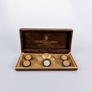 Op de afbeelding: Een Gucci horlogeset in een bruine fluwelen presentatiedoos. De set bevat een goudkleurig armbandhorloge met een verwisselbare ring, vergezeld van vier extra ronde ringen in de kleuren crème, bordeauxrood, marineblauw en donkerbruin.