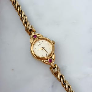 May include: A gold-tone Seiko wristwatch featuring a round white dial with gold hands and markers. The watch case is accented with two small round red stones at the top and bottom lugs. It includes a textured, twisted gold-tone metal link bracelet.