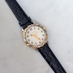 May include: A wristwatch with a black textured leather strap and a round face featuring a cream-colored dial. The dial displays black script numerals, gold-tone hands, and the text Chantal Thomass. The bezel has a scalloped gold and silver finish.