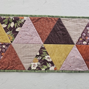 May include: A quilted table runner with a geometric design. The runner features a series of triangles in various colors and patterns, including floral, solid, and striped fabrics. The edges are finished with a green border.