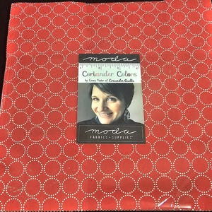 May include: A red fabric with a white dotted circle pattern. A card with the text "Coriander Colors" and a woman's portrait is centered on the fabric. The card also displays the "moda FABRICS + SUPPLIES" logo.