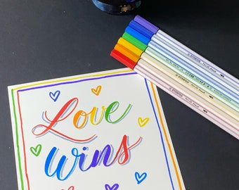 May include: A white card with the words "Love Wins" written in rainbow colors. The card is surrounded by a rainbow border. There are small hearts drawn around the words. A set of rainbow colored pens are lying on the card.