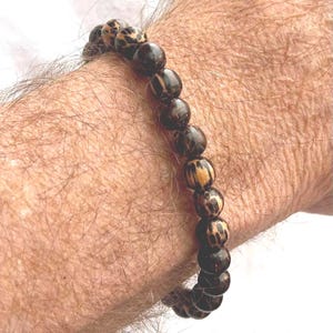 May include: A beaded bracelet featuring dark brown and tan spherical beads. The bracelet is made of natural stone and is worn on the wrist. The beads have a unique spotted pattern.