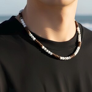 May include: A necklace made of white, brown, and dark brown beads. The beads are strung together to form a necklace. The necklace is worn against a black top. The background shows a blurred view of the sea.