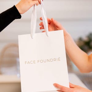 May include: A white shopping bag with a gold inscription that reads "FACE FOUNDRIE".