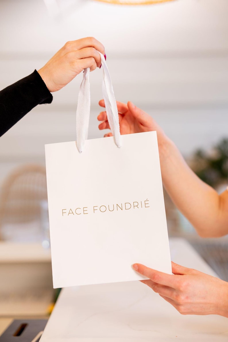 May include: A white shopping bag with a gold inscription that reads "FACE FOUNDRIE".
