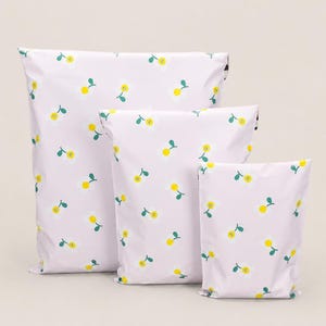 May include: Three white poly mailer shipping bags of varying sizes, each adorned with a repeating pattern of yellow smiling cherries with green stems. The bags are arranged in a stacked formation against a neutral background.