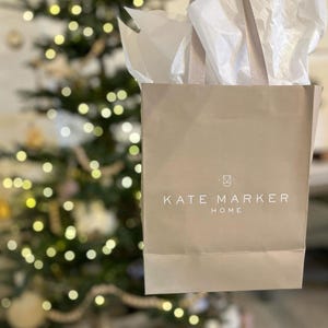 May include: A beige shopping bag with the text "KATE MARKER HOME" in white. The bag is filled with white tissue paper and has beige handles. A Christmas tree with lights is in the background.