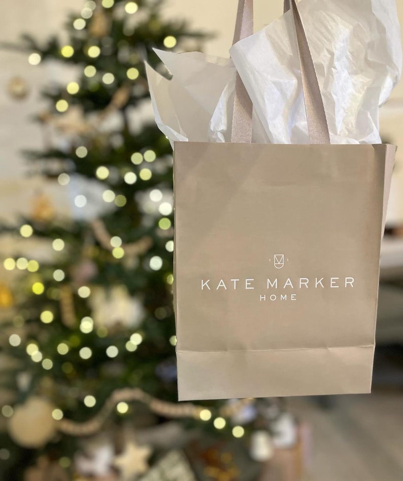 May include: A beige shopping bag with the text "KATE MARKER HOME" in white. The bag is filled with white tissue paper and has beige handles. A Christmas tree with lights is in the background.