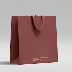 May include: A maroon paper shopping bag with the embossed word "abazo" on the front and a matching handle. The bag features the text "CANDLES & ACCESSORIES" and a short description. The bag is made of a textured paper.