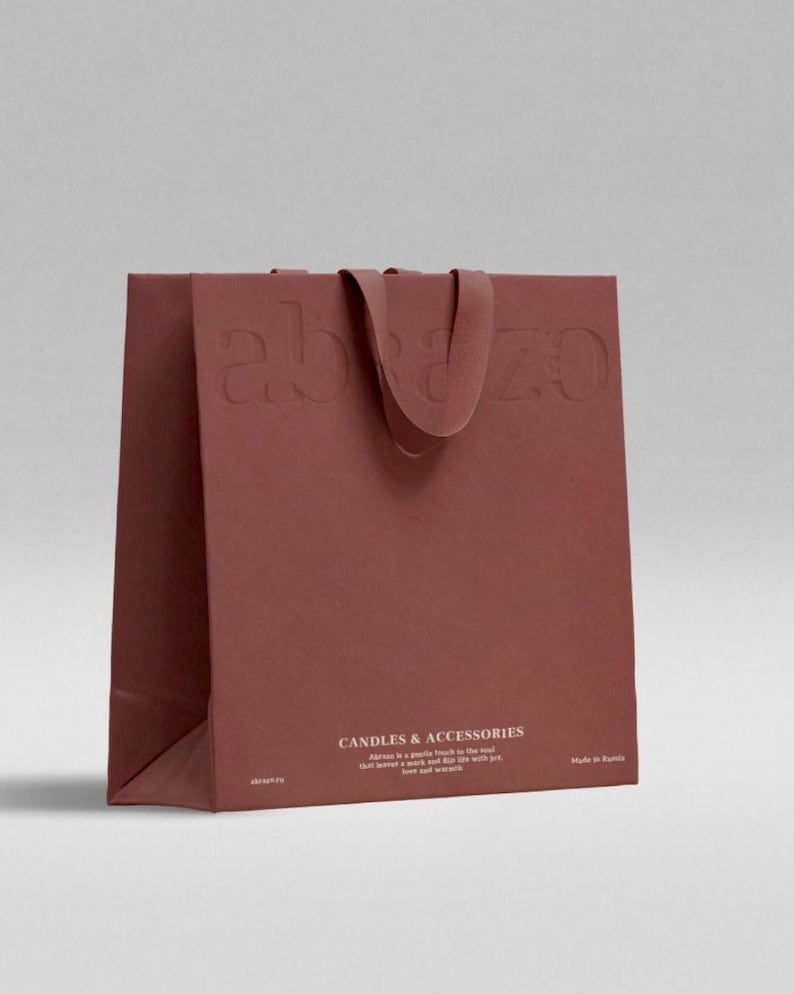 May include: A maroon paper shopping bag with the embossed word "abazo" on the front and a matching handle. The bag features the text "CANDLES & ACCESSORIES" and a short description. The bag is made of a textured paper.
