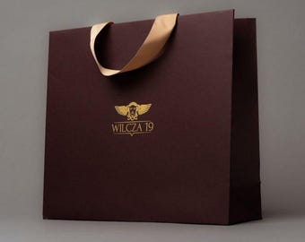 Puede incluir: Una bolsa de papel de color burdeos con un asa de cinta dorada y un logotipo dorado que dice "WILCZA 19" con un diseño de ángel alado.