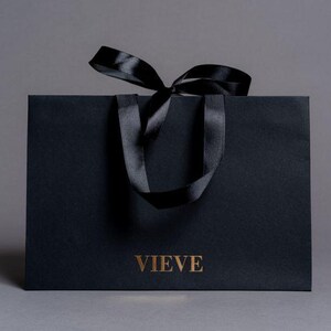 May include: A black paper shopping bag with a black satin ribbon bow and handles. The word "VIEVE" is printed in gold on the front of the bag. The bag is on a grey background.