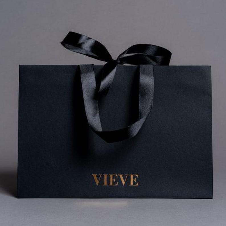 May include: A black paper shopping bag with a black satin ribbon bow and handles. The word "VIEVE" is printed in gold on the front of the bag. The bag is on a grey background.