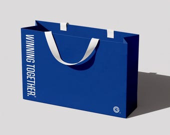 Puede incluir: Una bolsa de compras rectangular de color azul real con asas de cinta blanca. El texto WINNING TOGETHER está impreso verticalmente en blanco en el lado izquierdo, y un pequeño logotipo circular blanco con una G se encuentra abajo a la derecha.