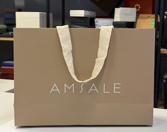 May include: A rectangular, light brown paper shopping bag with a cream-colored handle. The word "AMSALE" is printed in white on the front of the bag. The bag is sitting on a white surface.