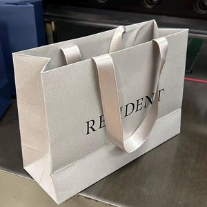May include: A light gray paper shopping bag with the word "RESIDENT" printed in black. The bag has matching satin ribbon handles. Another blue bag is visible in the background.