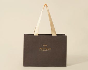 May include: A dark brown paper shopping bag with cream-colored handles. The bag features a gold logo and text that reads "pantala". The bag is sitting on a light beige background.