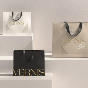 May include: Three shopping bags in cream, black, and tan, each with a black handle and the gold text "VERNIS". The bags are arranged on a white stepped surface, with a minimalist design.