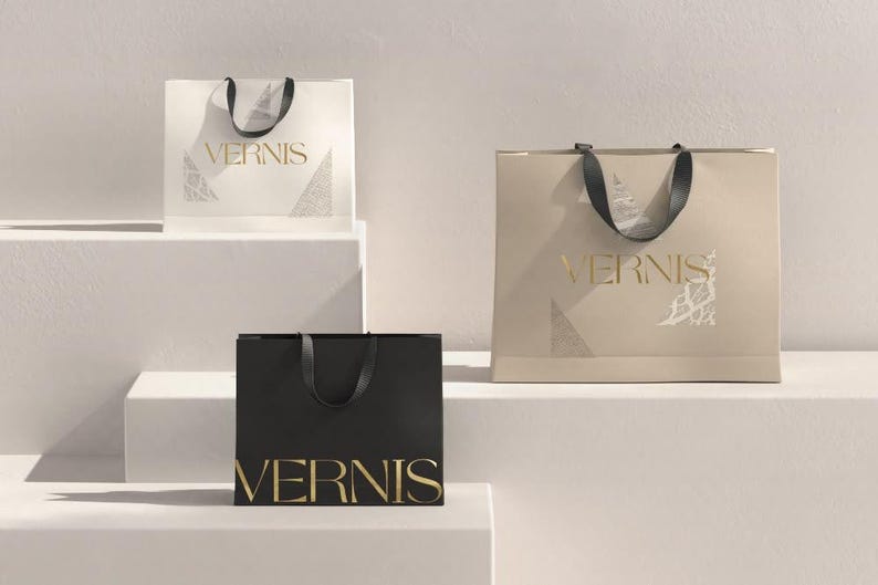 May include: Three shopping bags in cream, black, and tan, each with a black handle and the gold text "VERNIS". The bags are arranged on a white stepped surface, with a minimalist design.