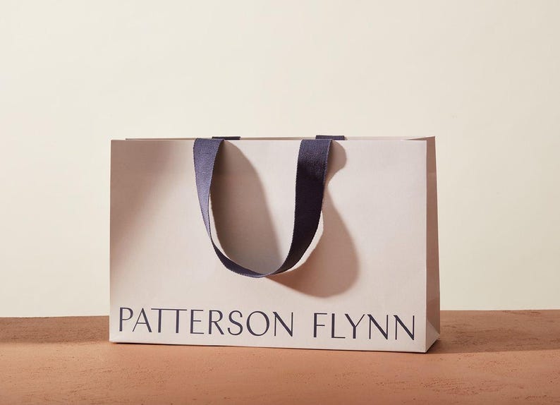 May include: A rectangular shopping bag with the text "PATTERSON FLYNN" printed in dark blue. The bag is light beige with dark blue handles. The bag is sitting on a brown surface.