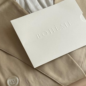 May include: A cream-colored card with the embossed word "BOHEME" rests on a tan fabric. The card has a curved edge and is slightly angled, revealing the texture of the fabric and a button.