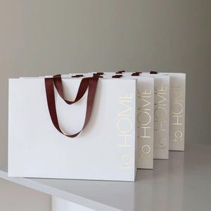 May include: Five white shopping bags with burgundy handles and gold lettering that reads "to HOME".