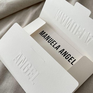 May include: A white box, open to reveal the text "MANUELA ANGEL" in black. The word "ANGEL" is embossed on the box, and "A HUMAN" is embossed on the top flap. The box rests on a neutral-toned surface.