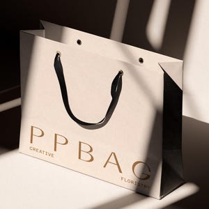 May include: A cream-colored shopping bag with black handles and the text "PPBAG CREATIVE FLORISTRY" in a gold color. The bag is sitting on a white surface with shadows.