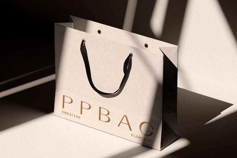 May include: A cream-colored shopping bag with black handles and the text "PPBAG CREATIVE FLORISTRY" in a gold color. The bag is sitting on a white surface with shadows.