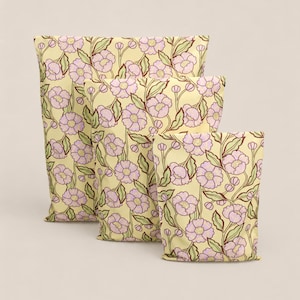 Puede incluir: Tres cojines decorativos con un estampado floral. Los cojines presentan un diseño repetido de flores lavanda, hojas verdes y tallos marrones sobre un fondo amarillo pálido. Los cojines están dispuestos en fila, con diferentes tamaños.