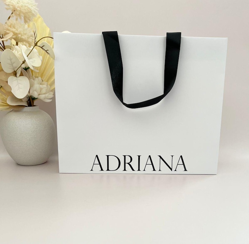 May include: A white shopping bag with black handles and the name "ADRIANA" printed in black. The bag is on a white surface. A vase with white flowers is in the background.