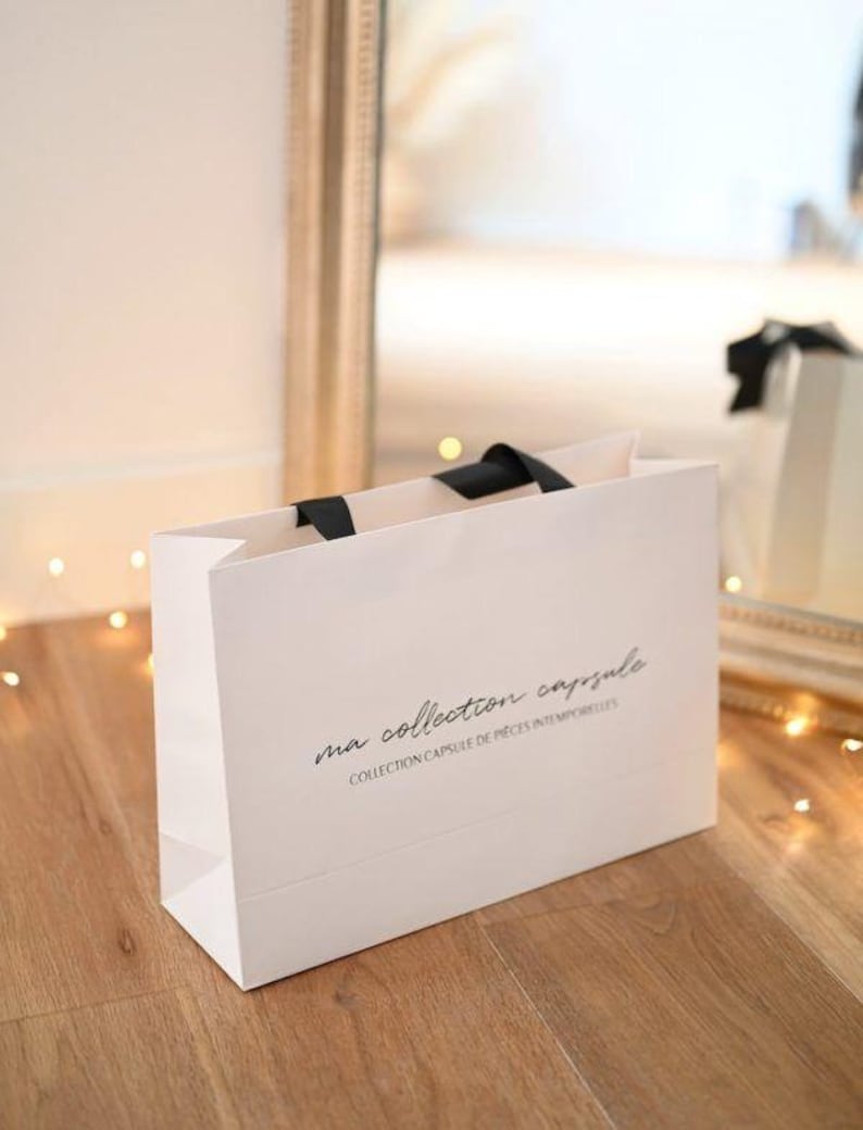 May include: A white shopping bag with black handles and the text "ma collection capsule" in a script font. The bag is on a wooden floor, with a mirror and lights in the background.