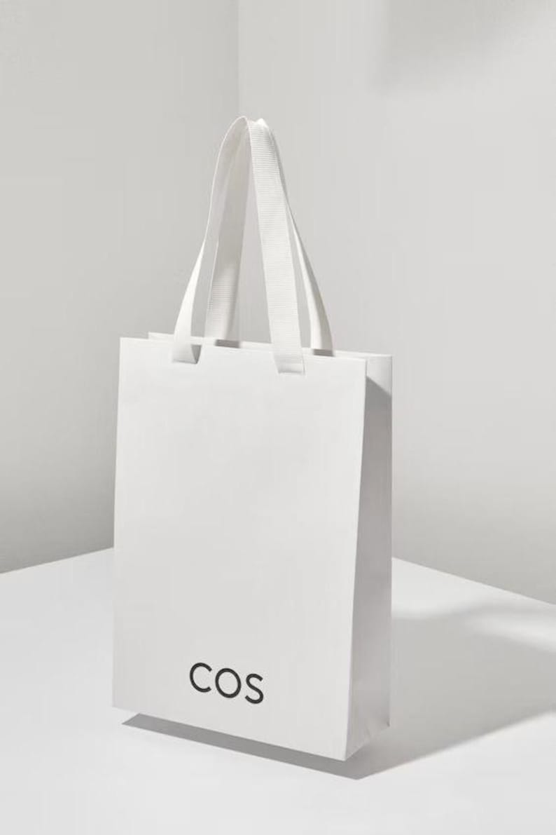 May include: A white shopping bag with the word "COS" printed in black on the front.