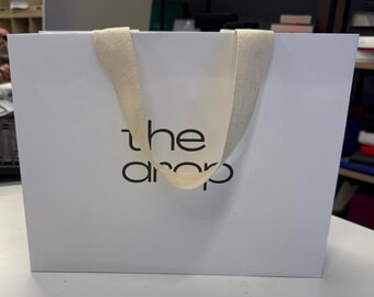 May include: A white paper shopping bag with the text "the drop" in black. The bag has a cream-colored fabric handle. The bag is sitting on a white surface.