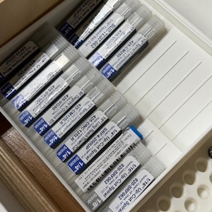 May include: A collection of router bits in a white plastic organizer. The bits are labeled with various specifications, including sizes like 1/4" and 1/8", and types such as "Spiral Compression" and "Up-Cut Spiral". The organizer is inside a wooden box.