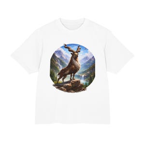 Puede incluir: Camiseta blanca con un gráfico circular de una cabra montés marrón con cuernos grandes y curvos. El gráfico muestra una cordillera, cascadas y un cielo azul. La camiseta está hecha de un material suave.