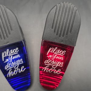 Puede incluir: Dos clips magnéticos de plástico, uno azul y otro rojo, con una sección superior negra con ranuras verticales. Los clips tienen el texto "place your design here" en escritura blanca. Útiles para sujetar papeles o notas.
