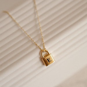 May include: A gold padlock necklace with the letter "E" engraved on the front. The necklace is on a white background.