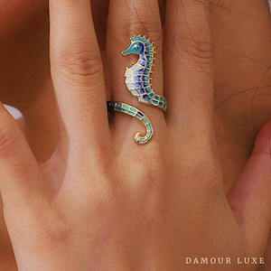 Può includere: Un anello color oro con un designo di cavalluccio marino in smalto blu e bianco. La coda del cavalluccio marino si avvolge attorno al dito.